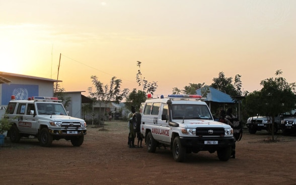 Two white off-road vehicles labelled "UN" and "Ambulance" are parked in a sandy square.